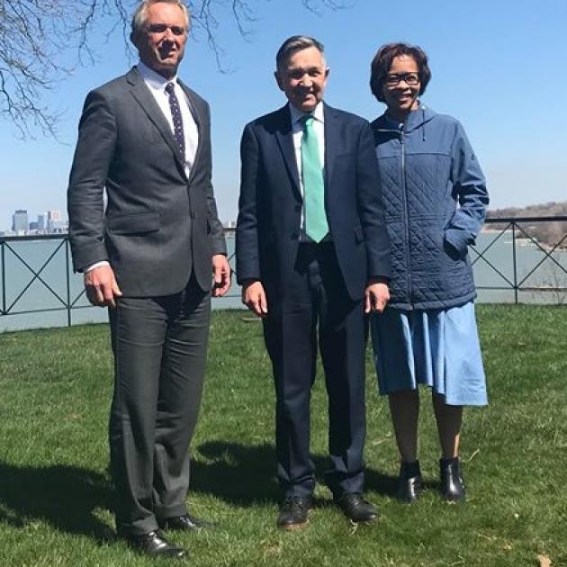 Three people standing outside on the grass with  body of water behind them, a tall white man in a suit with gray hair, and shorter white man in a suit and a black woman with glasses