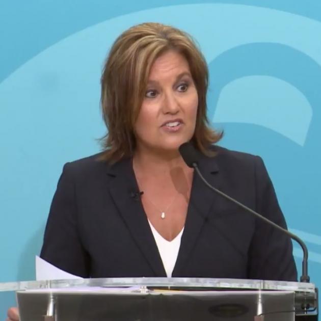White woman in a black suit with white shirt underneath and necklace with shoulder length brown hair standing at a mic with a blue background, her mouth in a form like she's talking