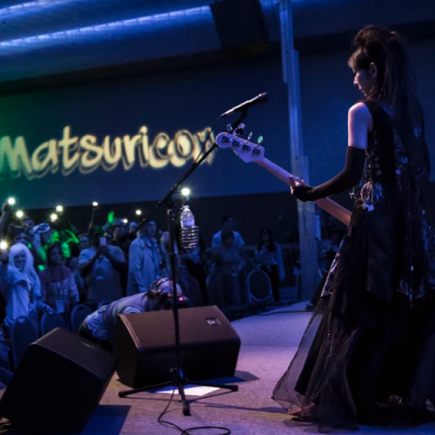 White woman in a Renaissance looking gown playing guitar on stage to a crowd standing under the sign Matsuricon