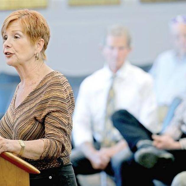 Middle-aged woman with red hair standing at a podium talking and people behind her in audience