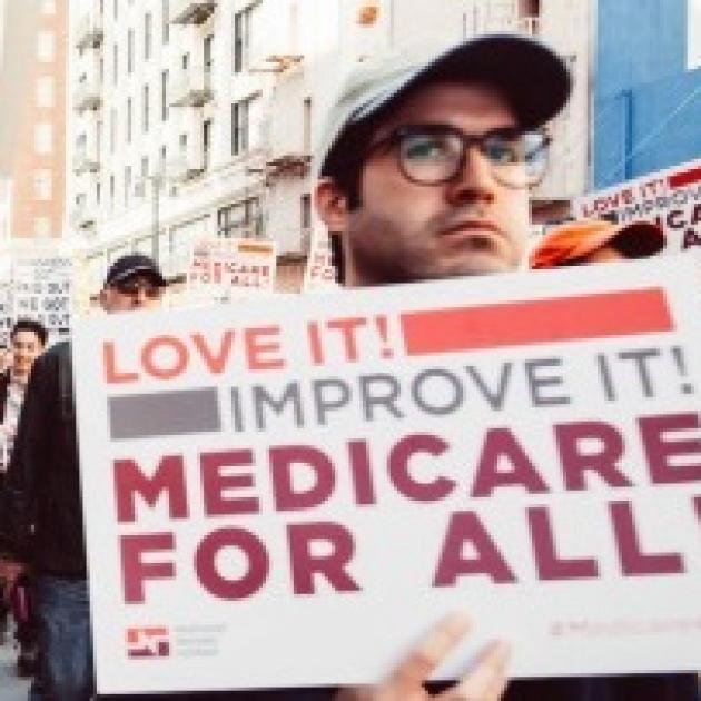 White man in a baseball cap and glasses holding a sign that says Love It Improve It Medicare for All