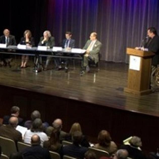 Panel of people on a stage with someone at the podium and an audience