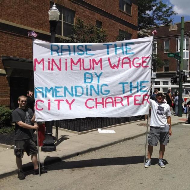 Two men holding sign reading Raise the MInimum wage by amending the city charter