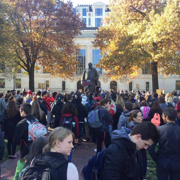 A crowd of people, mostly college students protesting around a statue with two trees on either side and a big building in the background