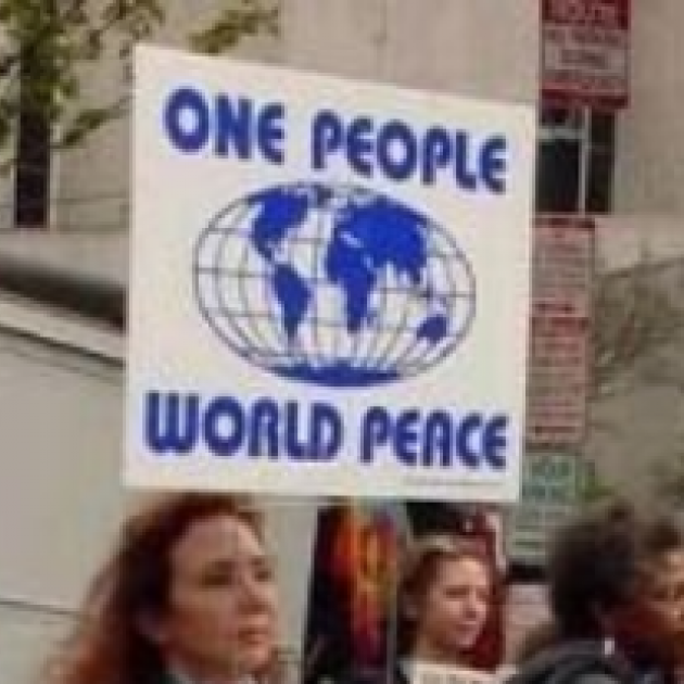 Woman outside with others holding a sign on a stick that has a globe in the middle and the words One People World Peace