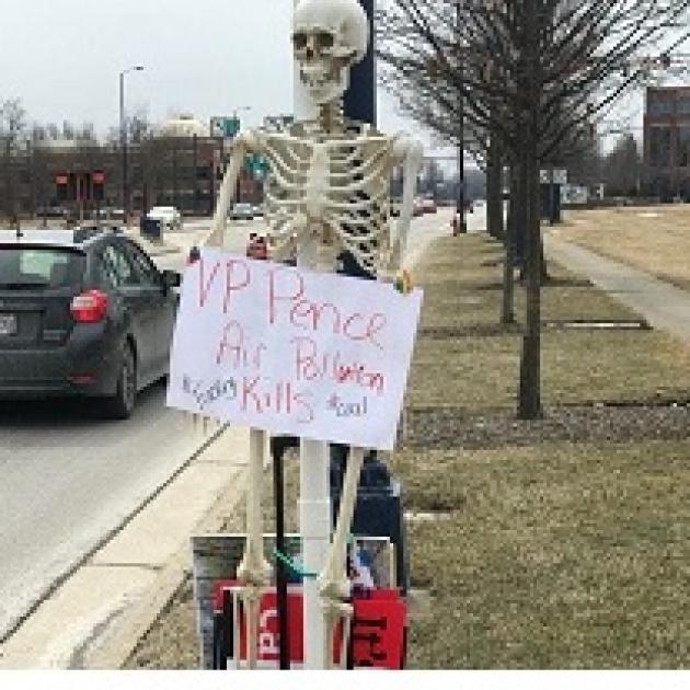 Skeleton leaning against a pole outside holding an anti-Pence sign
