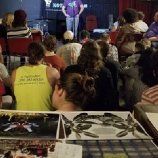 Lots of people with their backs to the camera sitting and watching someone standing at the mic, with art on a table in the foreground