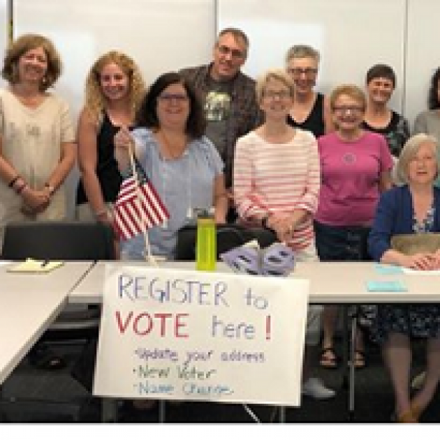 People posing standing behind a white table with a sign that says register to vote here