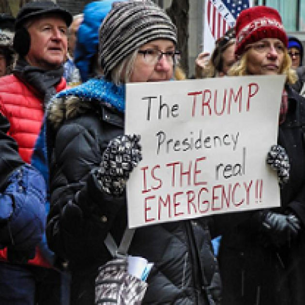 Woman in cold weather gear holding a sign at a protest outside that reads The Trump Presidency is the REAL emergency