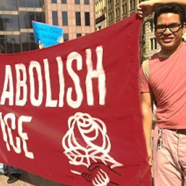 Young dark haired man with glasses holding a large red sign that says Abolish ICE with a DSA symbol of fist and rose