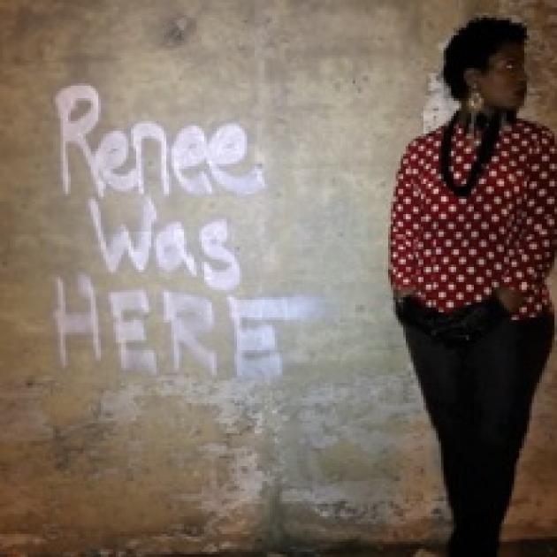 The words Renee was HERE spray painted on a brown wall and a black woman with short hair and a red and white shirt with a black necklace and black pants stands against the wall looking to the right
