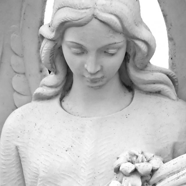 White statue of an angel, a woman with long flowing hair looking down and holding flowers in her arms