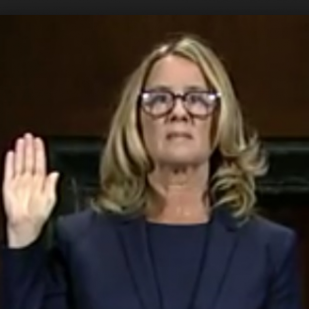 Blonde woman with glasses holding her hand up as if swearing to tell the truth