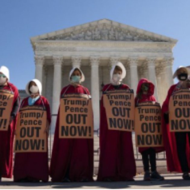 Proestors dressed like handmaidens outside a government building with signs saying Trump Pence Out Now