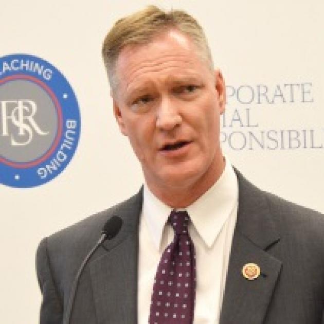 Thin white man in a suit speaking at a mic