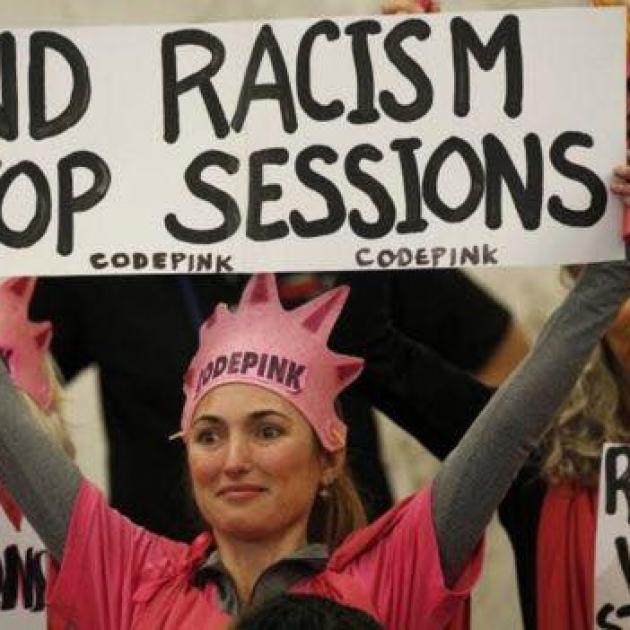 Blonde woman in pink with Code Pink crown holding a sign that says End racism Stop Sessions