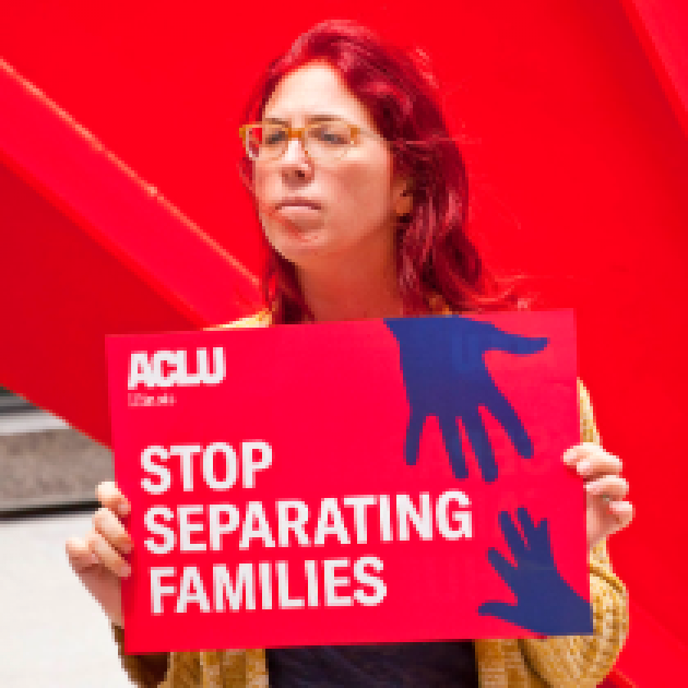 Woman with glasses and long red hair outside holding a sign that says Stop Separating Families