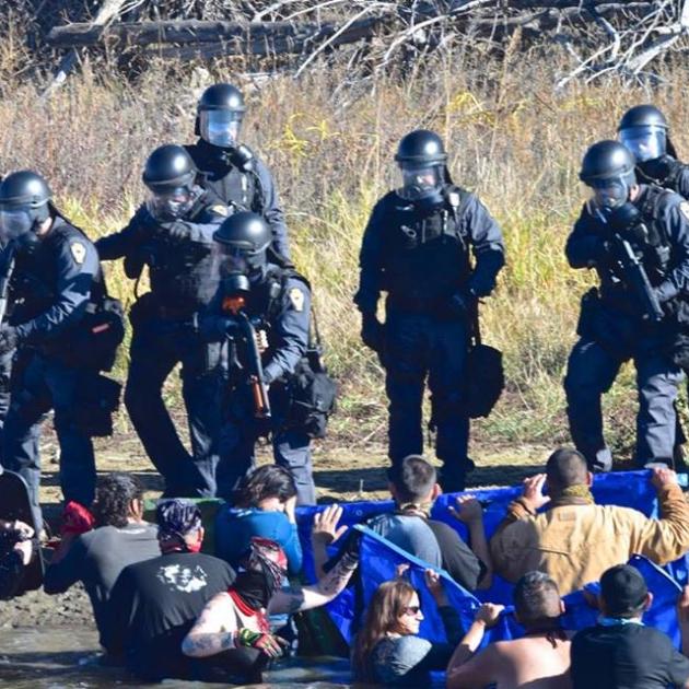 Photo of heavily armed soldier-looking men pointing guns at Native American protestors sitting on the ground