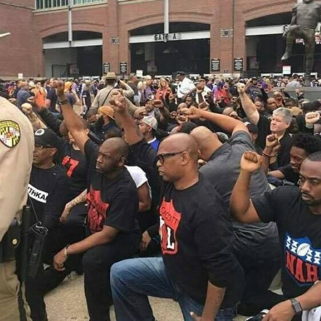 Lots of black men down on one knee with fists raised in the street with police man in foreground