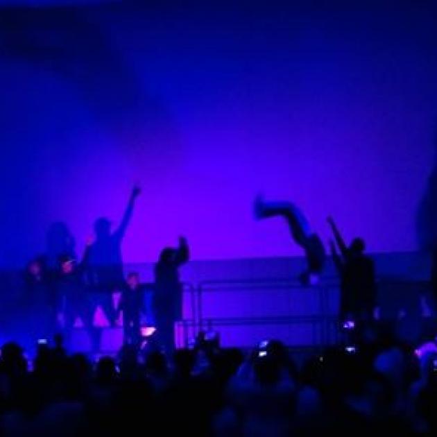 Silhouettes of people dancing and flipping in the air against a bright blue background on a stage in front of an audience