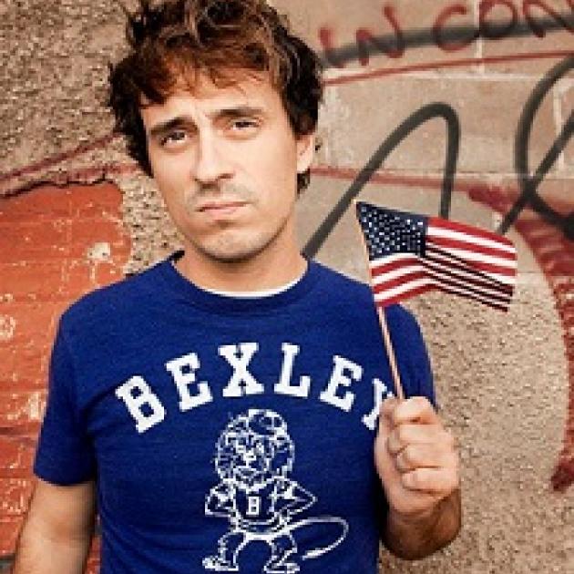 Young brown haired man wearing a blue t-shirt saying Bexley and waving a tiny flag in front of some kind of wall with words on it