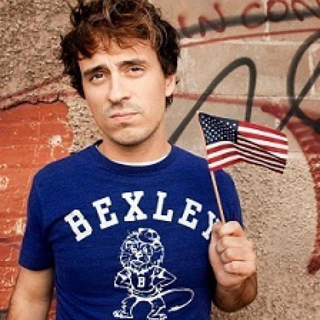 Young  white man  with tousled brown hair wearing a blue T-shirt that says BEXLEY in white letters and he's waving a very tiny flag and kind of sneering at the camera in a bored way