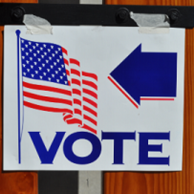 A white sign on a wood wall with a flag and an arrow pointing left and the word VOTE