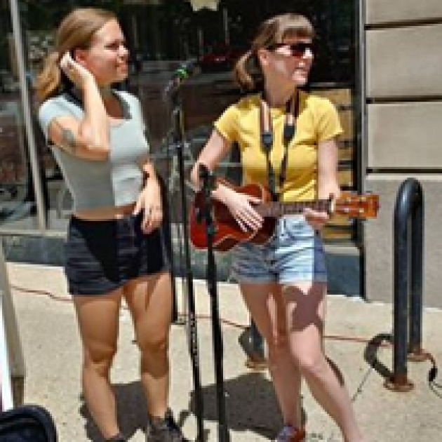 Two young white women wearing shorts standing outside by a microphone and one playing a guitar