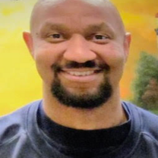 Bald black man with dark mustache and goatee, wearing blue shirt smiling