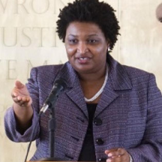 Black woman in a gray suit with a white necklace making a gesture while talking at a microphone