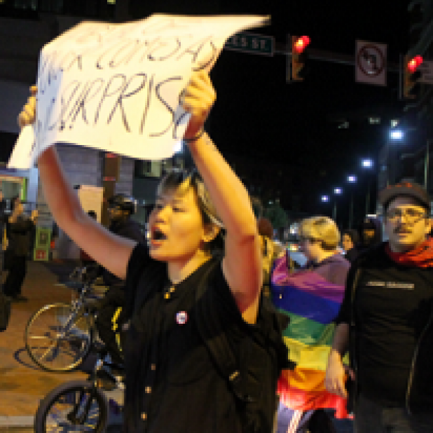 Young woman holding a protest sign above her head shouting and a person behind her in a rainbow flag and a guy next to her in a hat and glasses