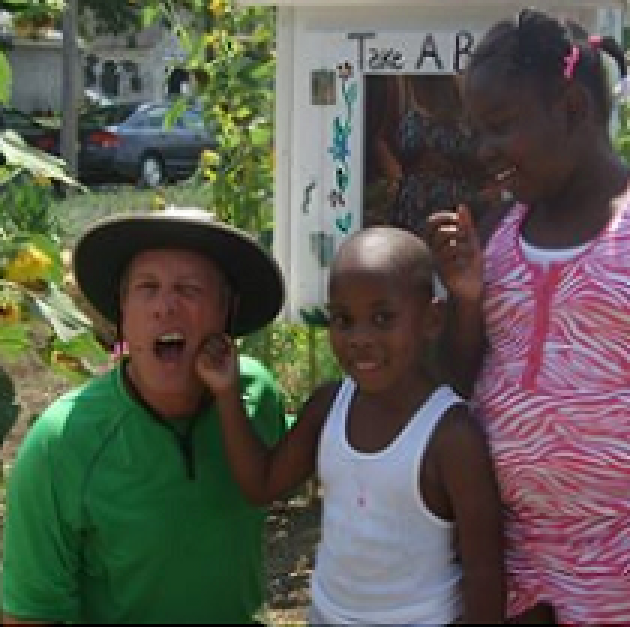 Two young black girls outside in a garden one touching the face of a white man bending down wearing a big hat