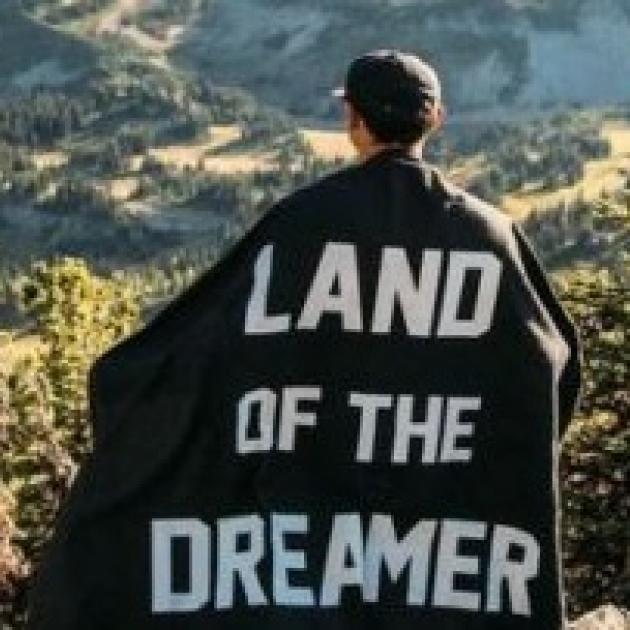 Man standing on a hill looking out with his back to the camera with a cape on with the words Land of the Dreamer
