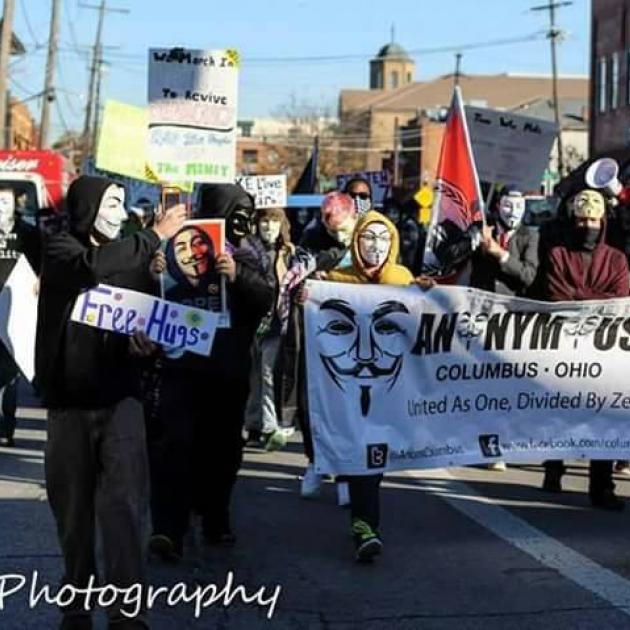 People marching wearing white and black Guy Fawkes masks carrying a long banner