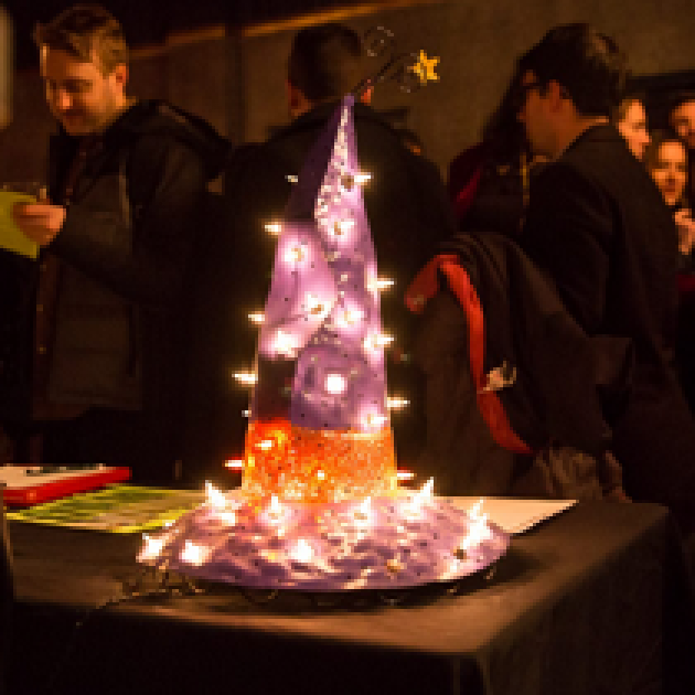 People at a party in the background and on a table in front a sparkly purple pointy hat with a wide brim and with an orange band around it