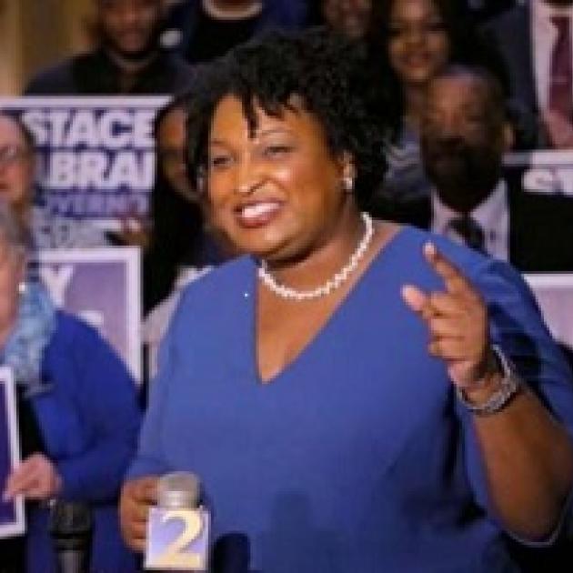 Older black woman with short curly black hair, white pearl necklace, blue dress, standing and smiling while gesturing in front of lots of people holding signs that say Stacey Abrams