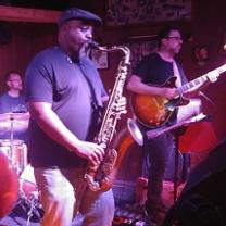 Heavy set black man in a cap playing the saxophone with other guys in a band onstage