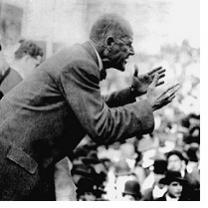 Black & white photo of a thin bald man sideways speaking and gesturing with arms outstretched to a crowd of people