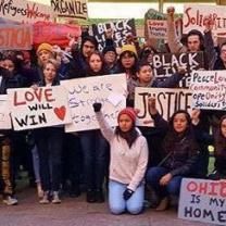 Many people posing in rows outside wearing winter clothes holding signs that say Live Will Win, Refugees Welcome, Justice, Ohio is my Home, Solidarity, Organize, and a woman in front on her knees with a fist in the air