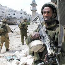 Foreground has a dark skinned man holding as assault rifle in the background two military men with rifles walking toward a building