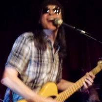 Man with long brown hair in plaid shirt playing a guitar and singing into a mic