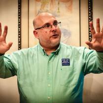 White middle aged guy with glasses holding his arms in the air with fingers spread on hands while he talks and wears an Abolish the Death Penalty button