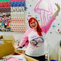 Woman with red hair and pink glasses holding up her hand like waving hello in a room with a lot of arts and crafts supplies
