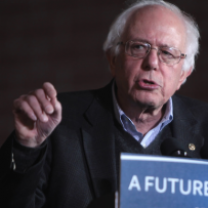 Older white man with white hair and wire rimmed glasses gesturing and speaking at a podium