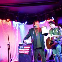 Two men on a stage, one with a guitar, one with a plaque in his hand
