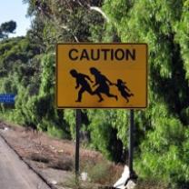 Yellow sign by the road and a hill with silhouettes of a family holding hands and running and the words CAUTION