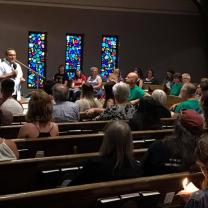 Man in white shirt talking into a mic at the far left side and a congregation of people sitting in pews and three stained glass windows in the back