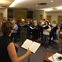 Lots of women in a half circle singing with one woman conducting