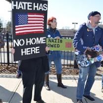 Someone holding a sign with a US flag on it on black background saying Hate has no home here, a guy playing a guitar that's covered with stickers with political sayings and a woman with red hair behind in the middle with a sign against Nazis