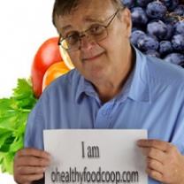 Older white man with glasses in front of fruits and vegetables holding a sign that reads I am healthyfoods.com